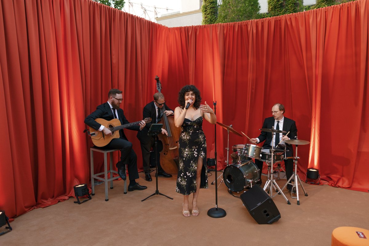 A jazz band, consisting of a female vocalist, guitarist, bassist, and drummer, performs on a stage with a vibrant red cu