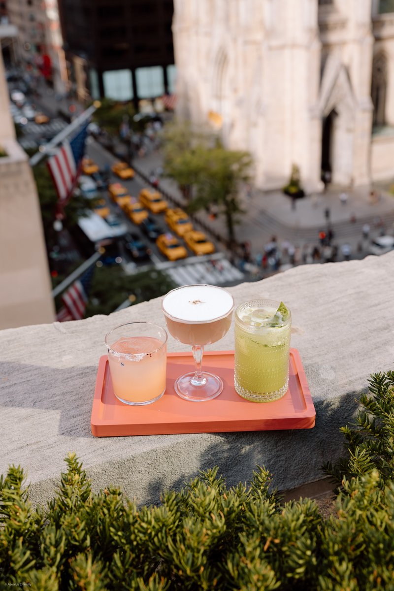 Three colorful cocktails are presented on a coral-colored tray, set on a stone ledge with lush green foliage in the fore