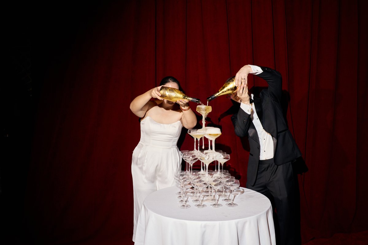 Two individuals, one in a white jumpsuit and the other in a black tuxedo, are actively pouring champagne from golden bot
