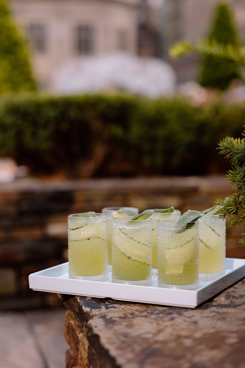 A tray of refreshing green cocktails, garnished with cucumber slices and basil leaves, sits on a rustic stone wall outdo