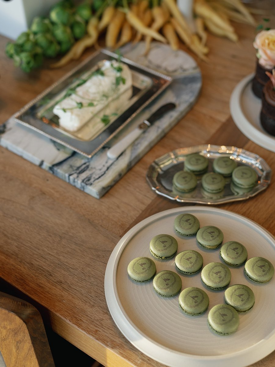 A close-up shot of a rustic wooden table laden with various food items. Green macarons with a custom logo are neatly arr
