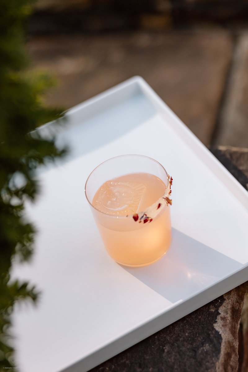 A refreshing pale pink cocktail, garnished with a citrus peel and dried flower petals, sits on a clean white serving tra