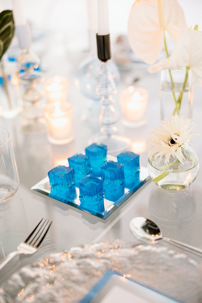A close-up shot of an elegantly set table featuring vibrant blue gelatin cubes on a mirrored tray. The setting includes 