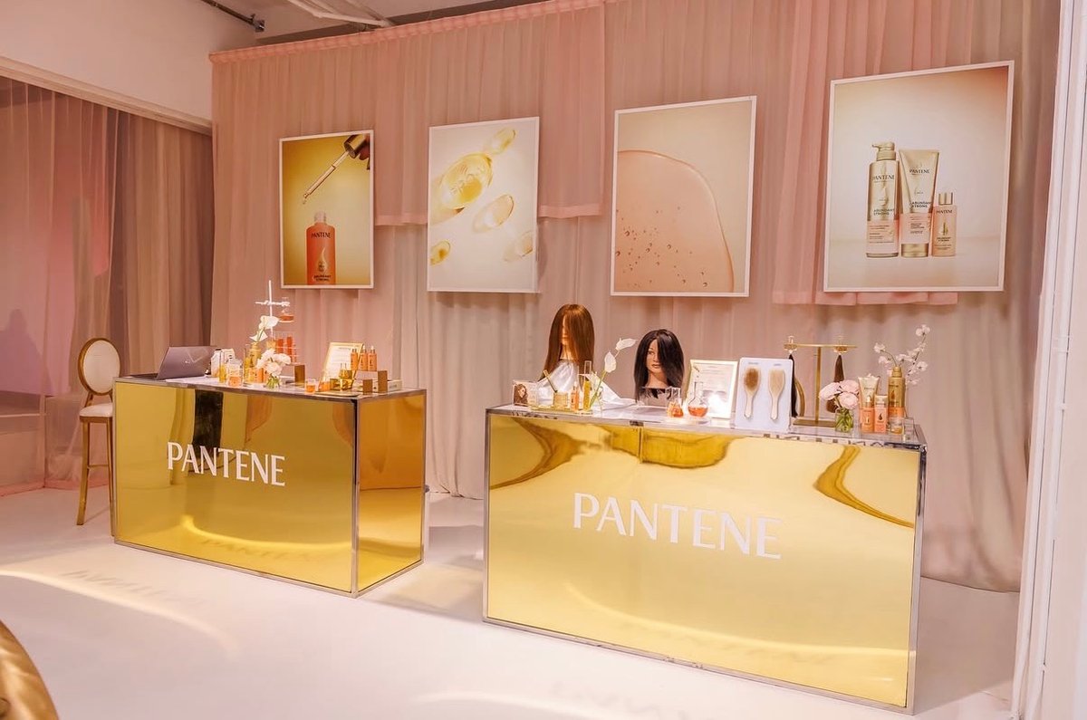 Two golden, mirrored display stations with 'PANTENE' branding are set up against a backdrop of sheer blush pink curtains