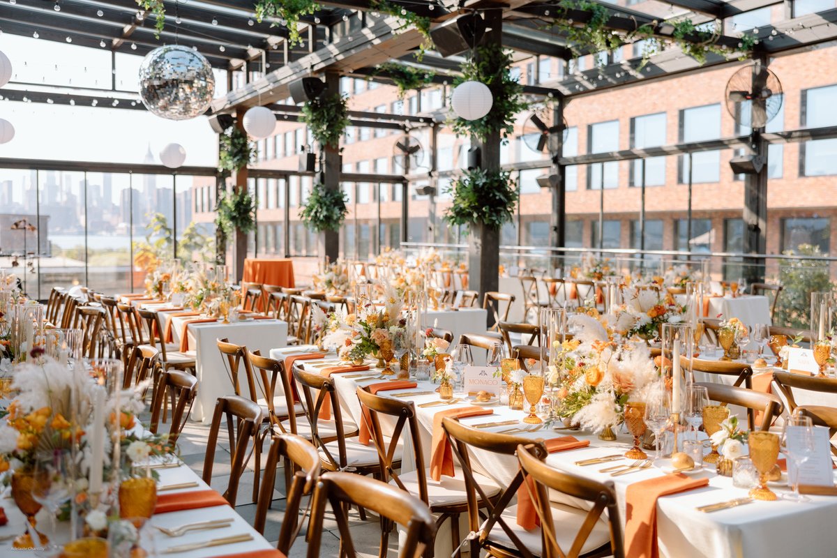 A beautifully set rooftop event space features long dining tables adorned with vibrant orange and white floral arrangeme
