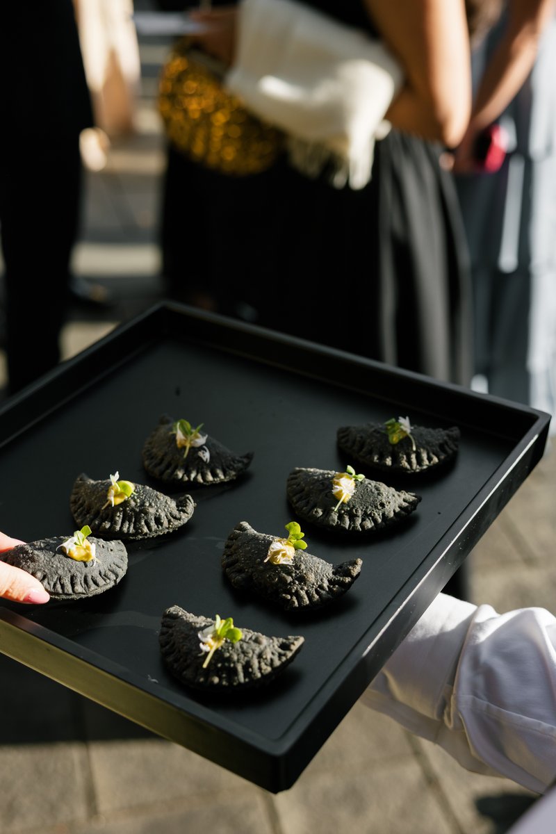 A server presents a tray of unique black empanada-style appetizers, each garnished with a dollop of yellow sauce and mic
