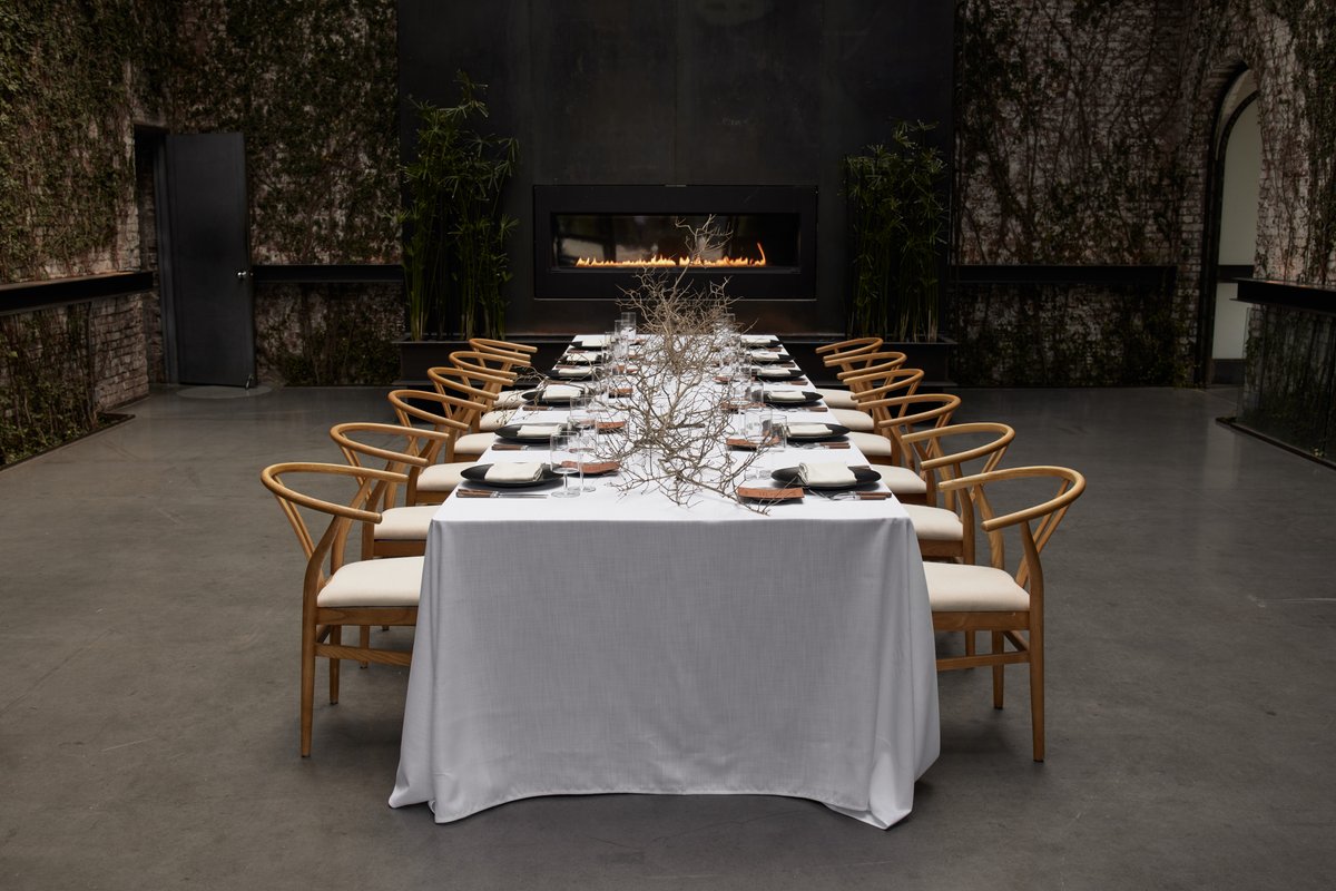 A long, elegantly set dining table with light wood chairs and a white tablecloth is centered in a unique event venue. Th