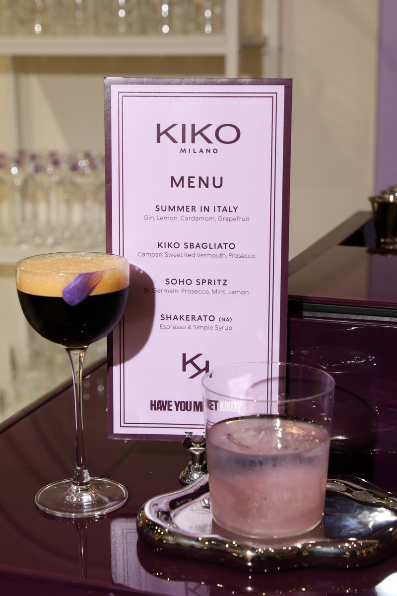 Two stylish cocktails are presented on a glossy purple surface alongside a Kiko Milano branded menu. One drink is dark w