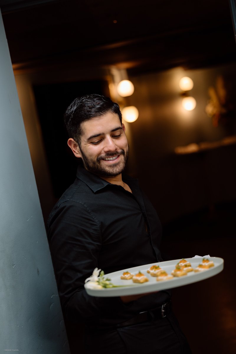 A smiling server, dressed in a black shirt, holds a tray of appetizers, looking down at the food with a pleasant express