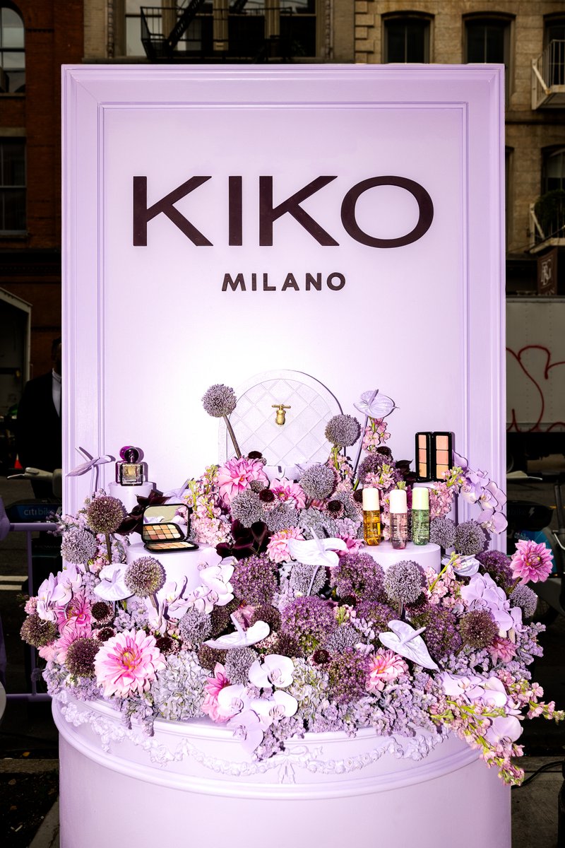 A vibrant Kiko Milano product display features an abundance of purple and pink flowers, showcasing various makeup items 