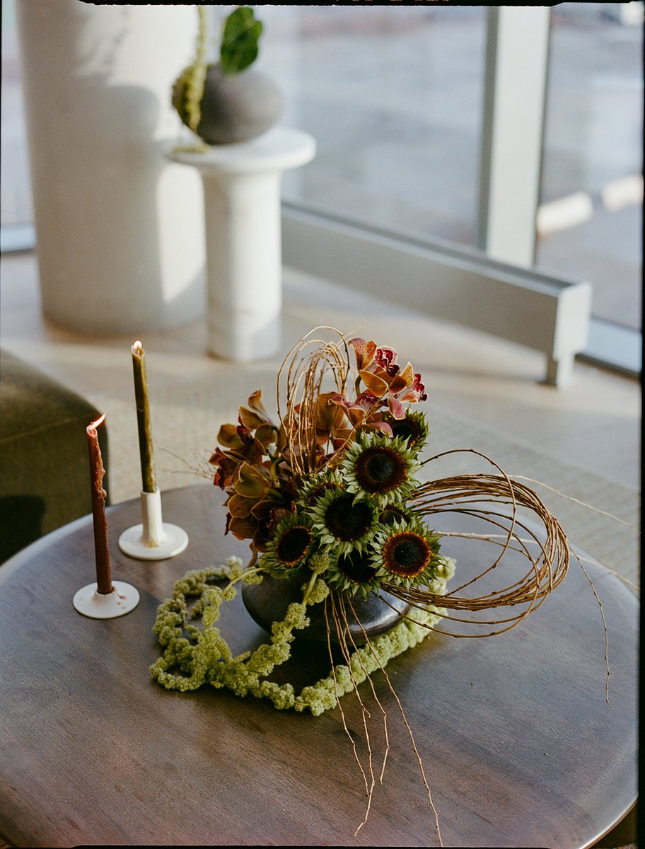 An elegant floral centerpiece featuring sunflowers, orchids, and decorative branches sits on a dark wooden table. Two li