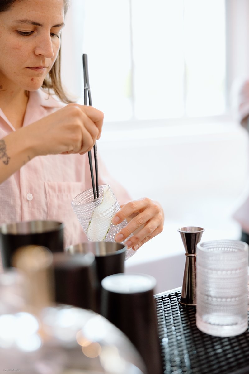 A bartender, dressed in a light pink shirt, is meticulously garnishing a textured glass with a cucumber slice using blac