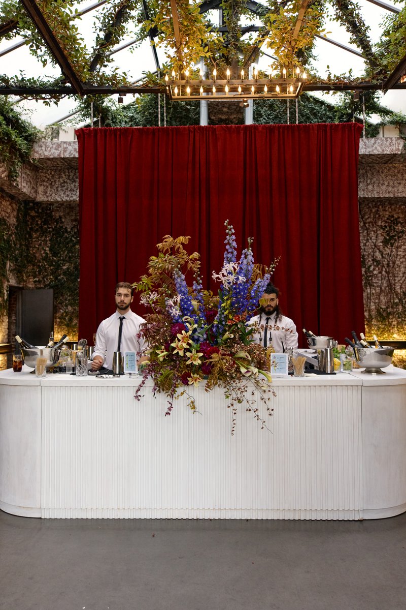 Two bartenders stand behind a stylish white bar adorned with a large, vibrant floral arrangement, set against a rich red