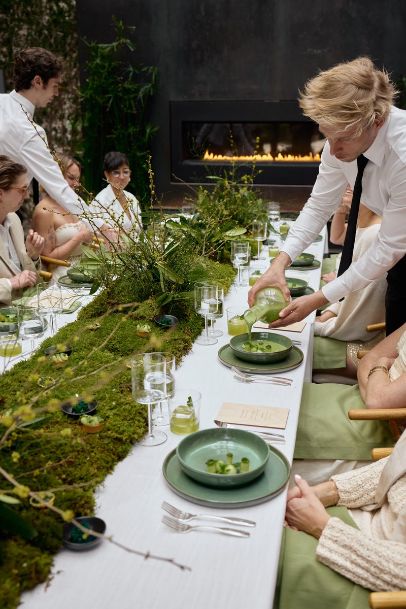 Servers attend to guests seated at a long, elegantly decorated table, featuring a lush moss and botanical runner. One se