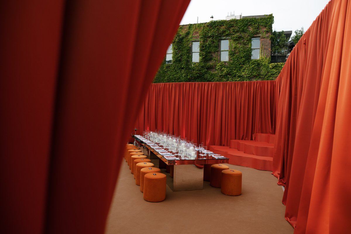 A vibrant outdoor event space is set with a long, reflective dining table surrounded by orange cylindrical stools. Drama