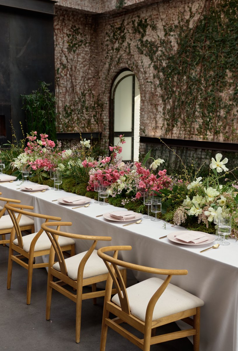 A beautifully set long dining table features an elaborate centerpiece of pink and white flowers nestled in green moss, a