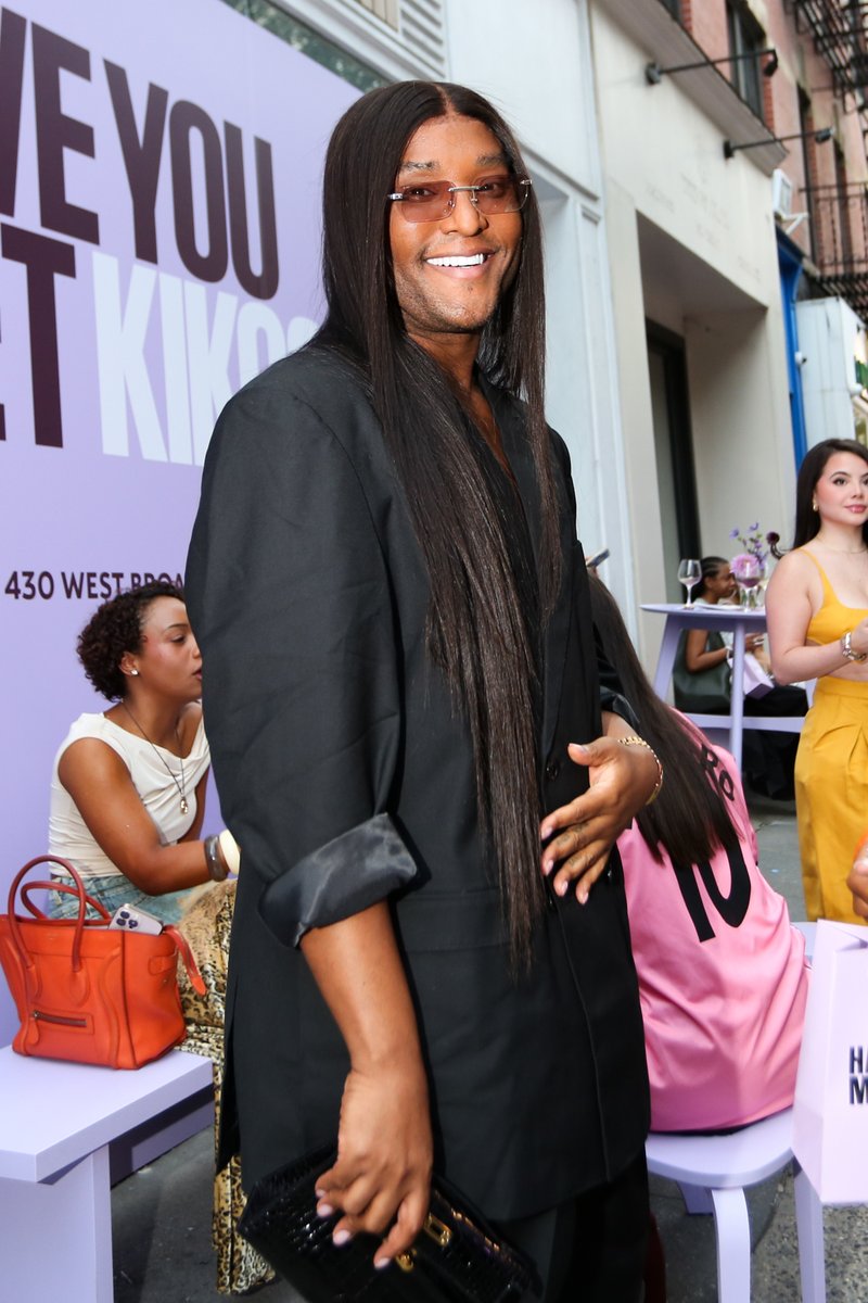 A person with long dark hair and sunglasses smiles brightly at the camera, holding a black clutch. They are wearing a bl