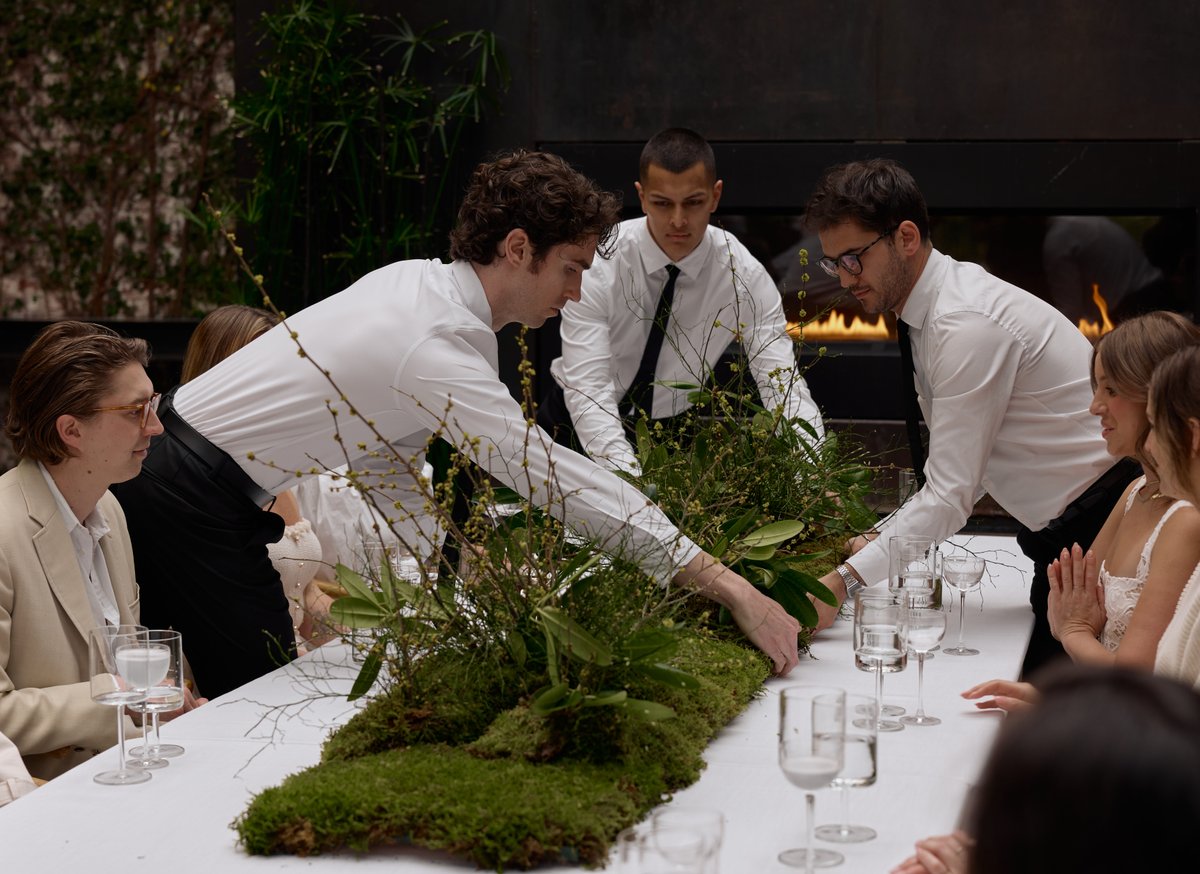 Three servers in white shirts and black ties are meticulously arranging a long, lush green moss and plant centerpiece on