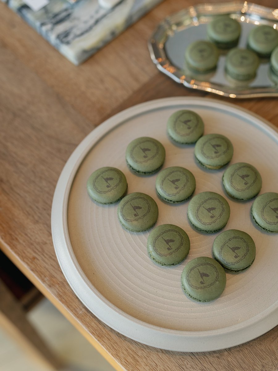 A collection of custom green macarons, each adorned with a golf flag logo, is elegantly arranged on a round, textured pl