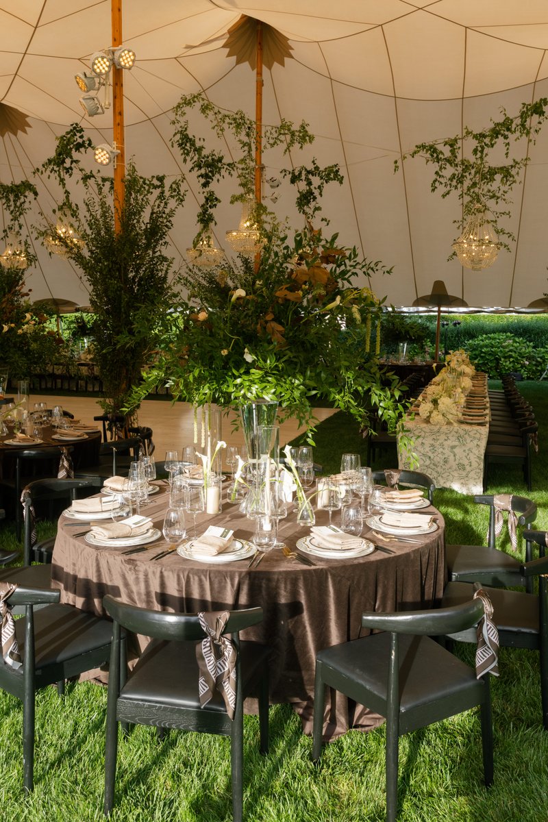 A beautifully set round dining table is featured under a large white tent, surrounded by vibrant green grass. The table 