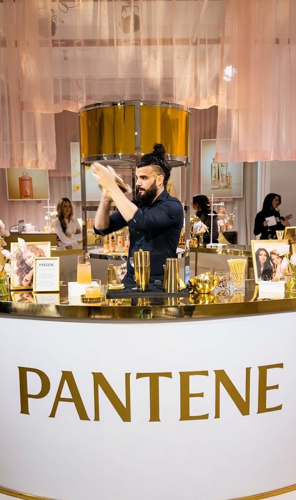 A male bartender with a beard and man bun is actively shaking a cocktail at a sleek, branded bar. The bar features gold 