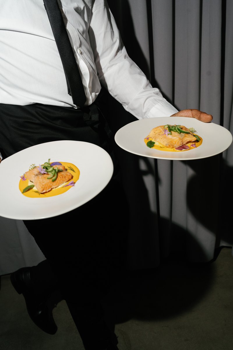 A server, dressed in a white shirt and black apron, holds two elegantly plated dishes, likely salmon with a vibrant yell