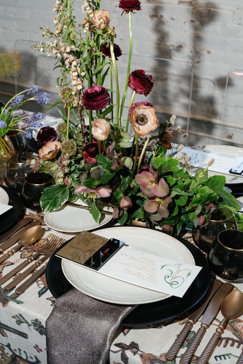 A beautifully arranged table setting features a vibrant floral centerpiece with deep red, blush, and cream flowers, surr