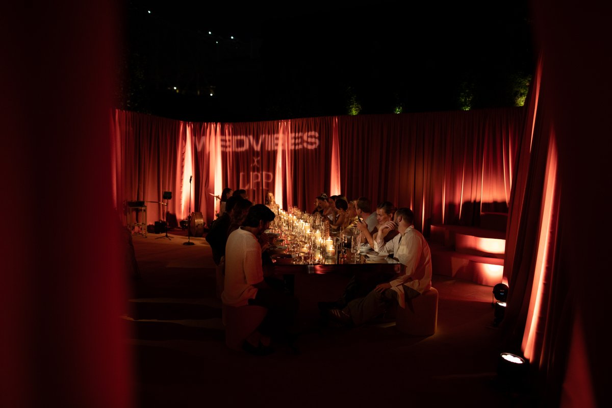 A group of guests are seated around a long, candlelit table in a dimly lit room, surrounded by dramatic red drapes. The 
