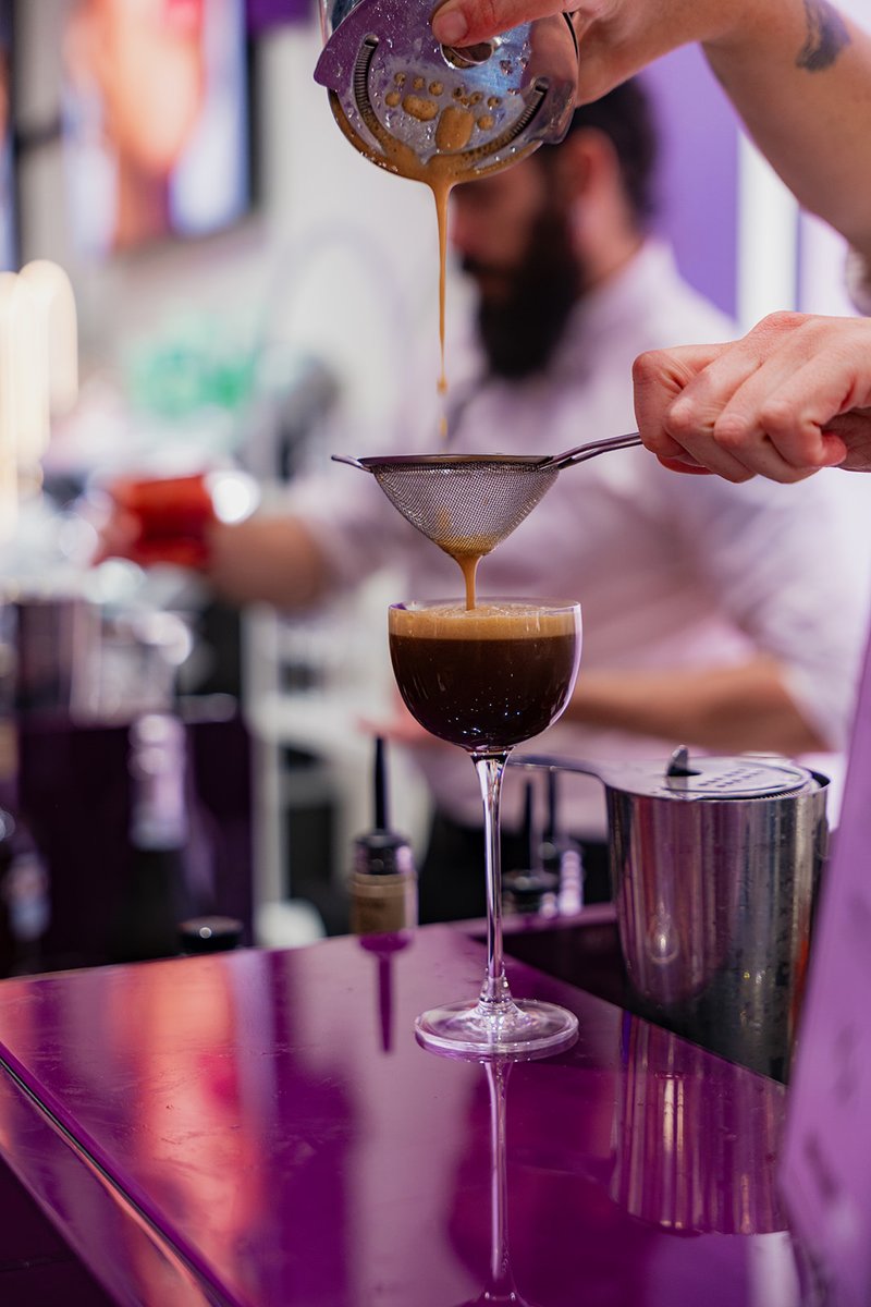 A bartender's hands are actively pouring a dark, frothy drink through a fine-mesh strainer into an elegant coupe glass. 