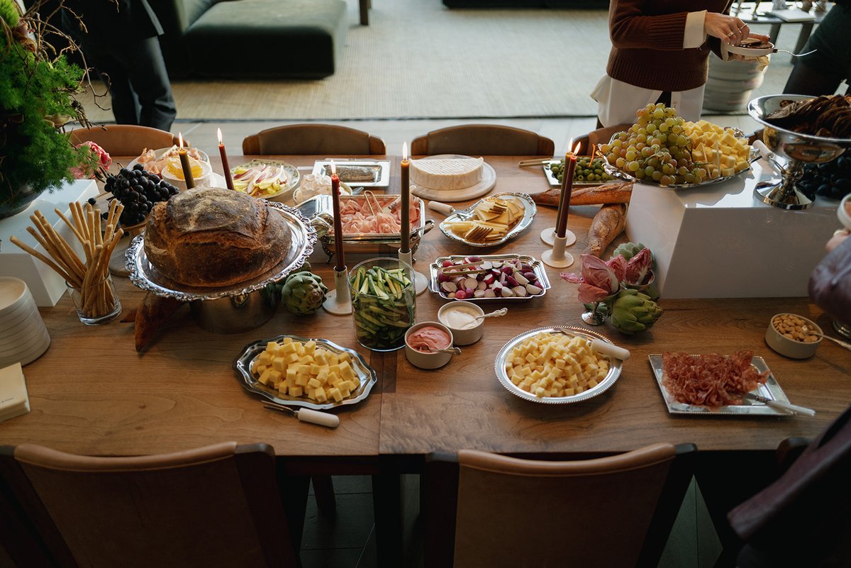 A beautifully arranged buffet table showcases a variety of gourmet cheeses, cured meats, fresh bread, and crudités. Lit 