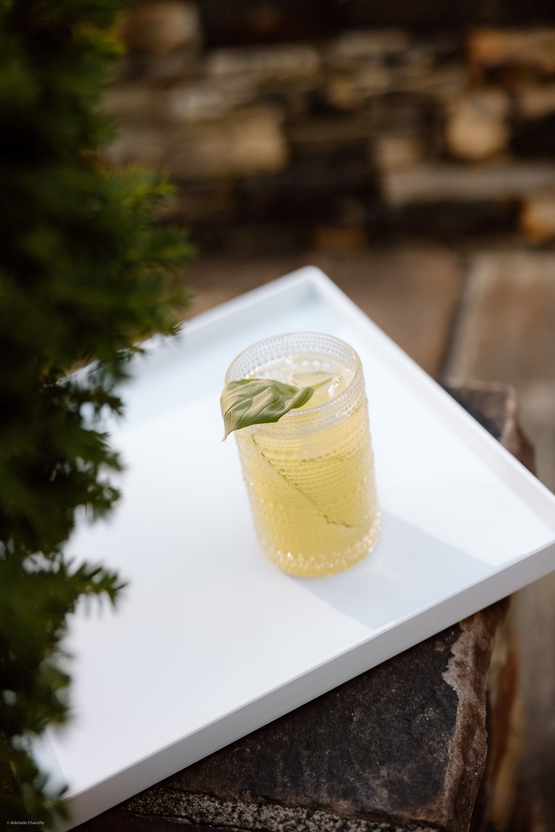 A vibrant yellow cocktail, garnished with a fresh green leaf, is presented in a textured glass on a clean white serving 