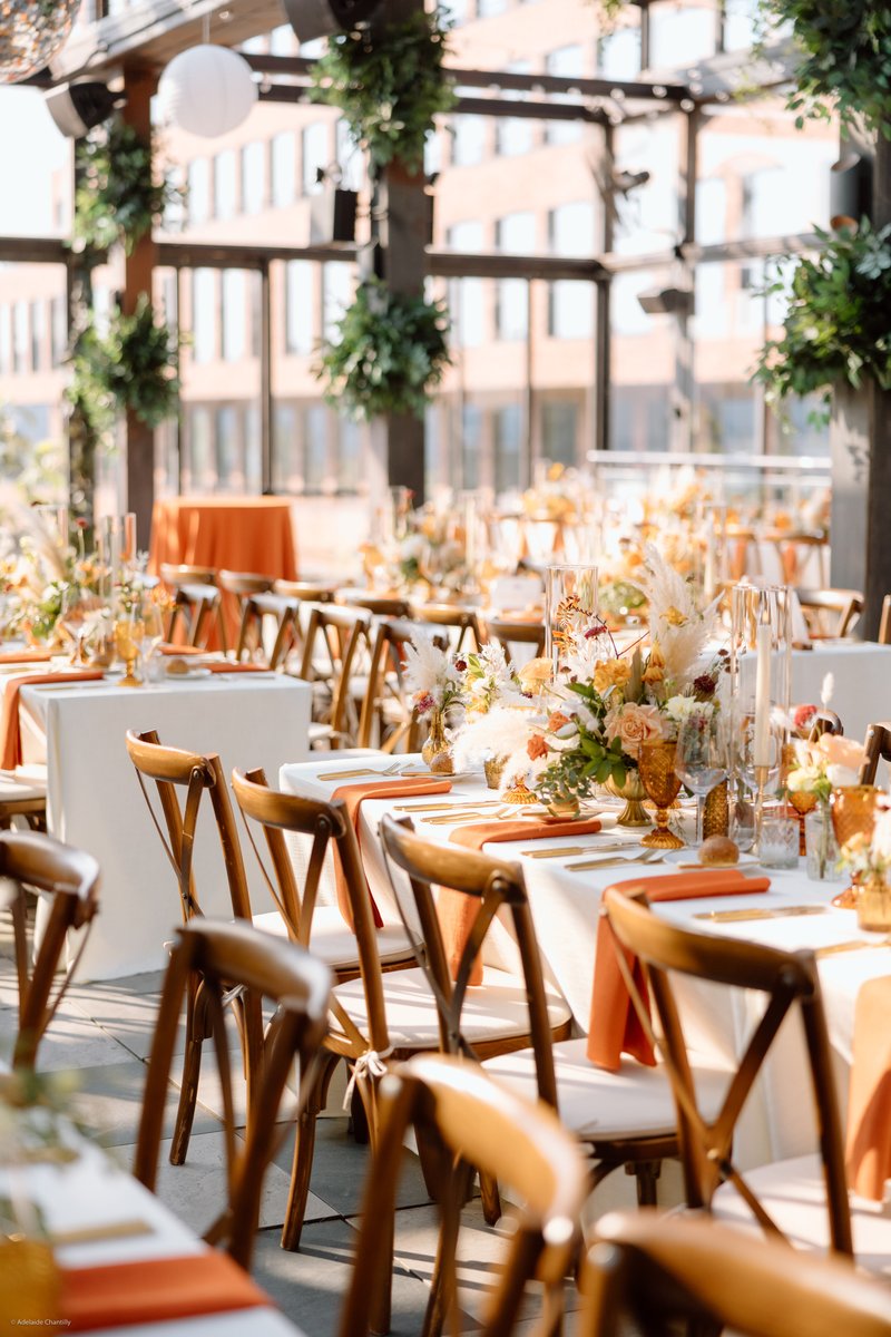 A beautifully set outdoor or rooftop event space features long tables adorned with white tablecloths and burnt orange ru