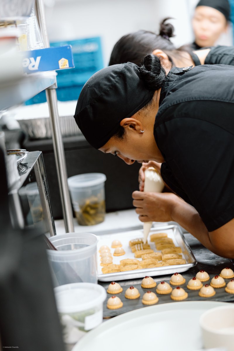 A chef in a black uniform and hairnet meticulously pipes a creamy topping onto small, rectangular desserts arranged on a