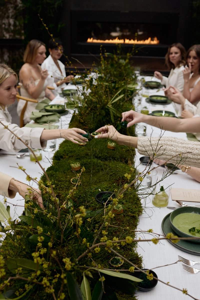 Guests are seated at a long, elegantly set table adorned with a lush, mossy runner and green foliage. Several individual