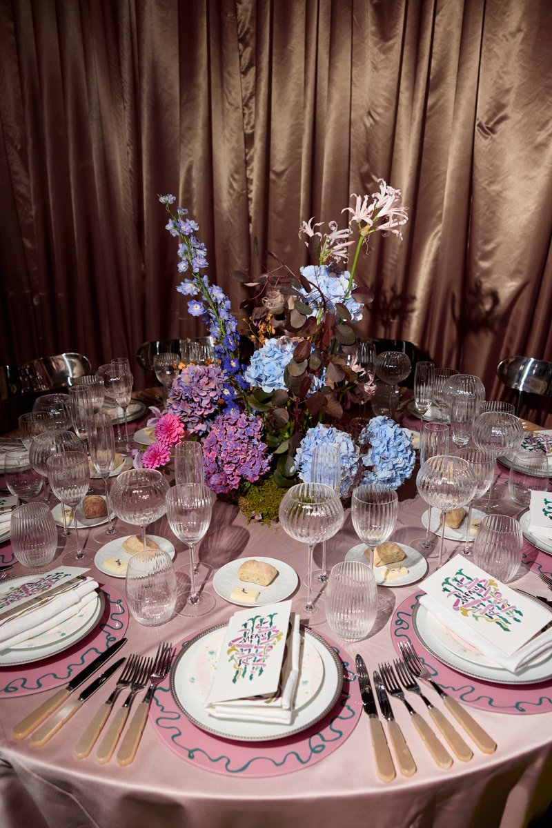 A beautifully set dining table features an elaborate floral centerpiece with blue, purple, and pink hydrangeas and other