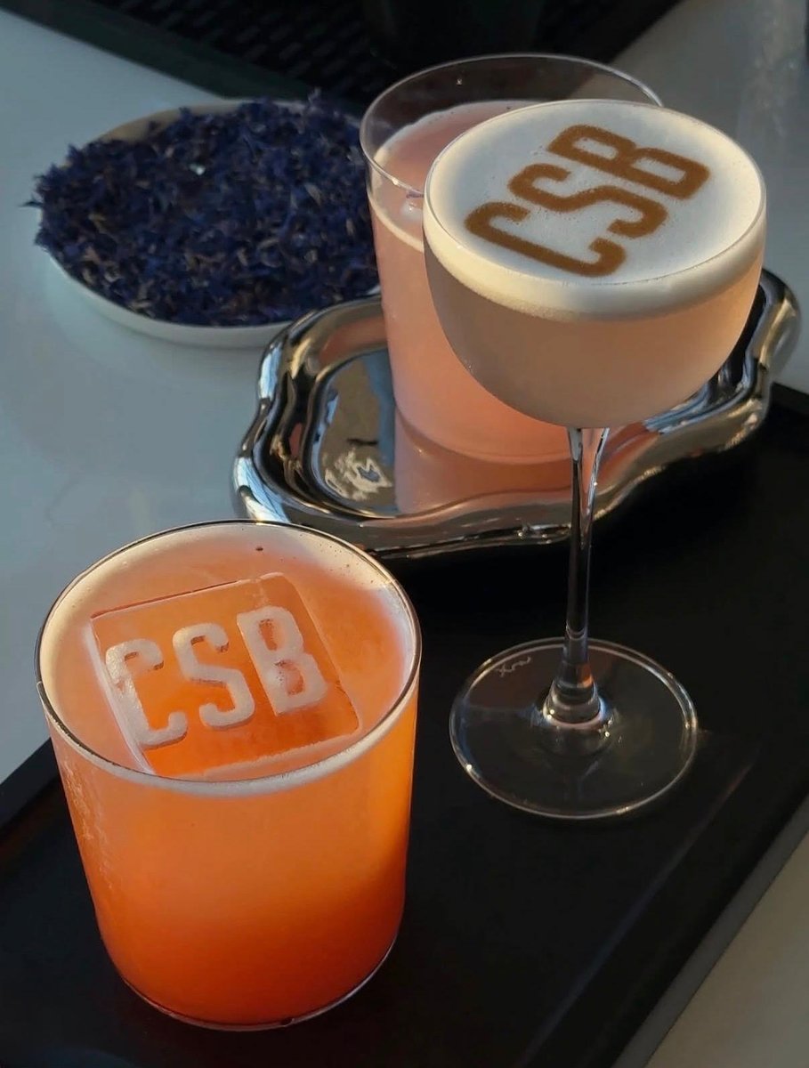This close-up shot features two custom cocktails, one with 'CSB' branded foam art and another with a 'CSB' ice cube, pre