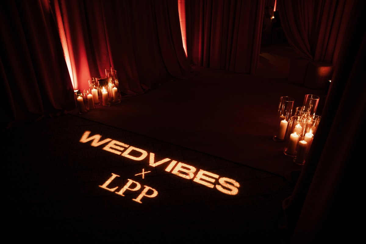 A dimly lit event space features rich red drapes, clusters of pillar candles in glass vases, and a projected "WEDVIBES x