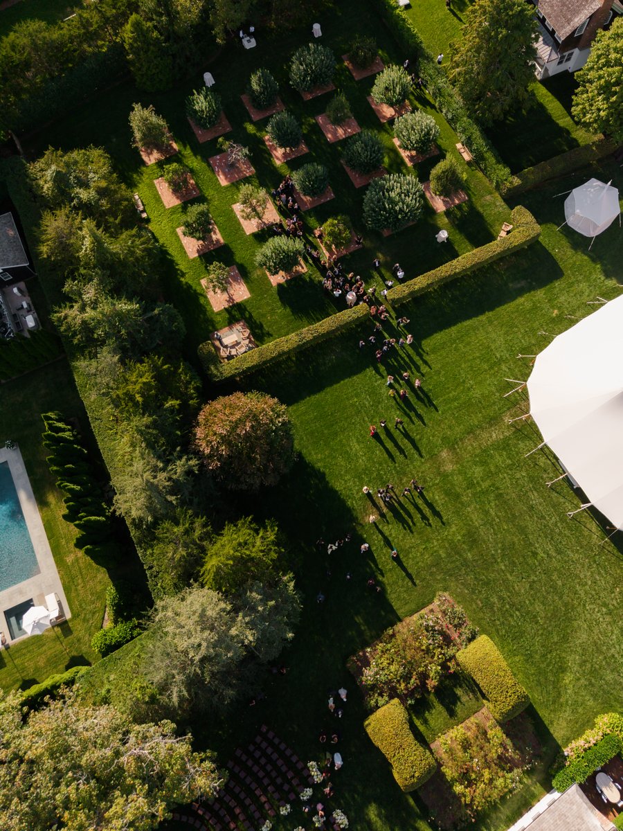 An aerial view captures a sprawling outdoor event space, featuring manicured lawns, a formal garden with square beds, an