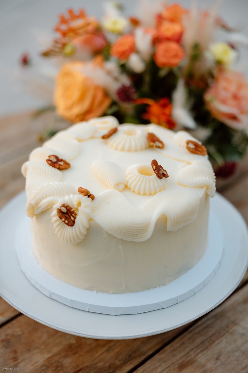 A beautifully decorated white cake with piped frosting and pecan halves sits on a white plate, placed on a rustic wooden