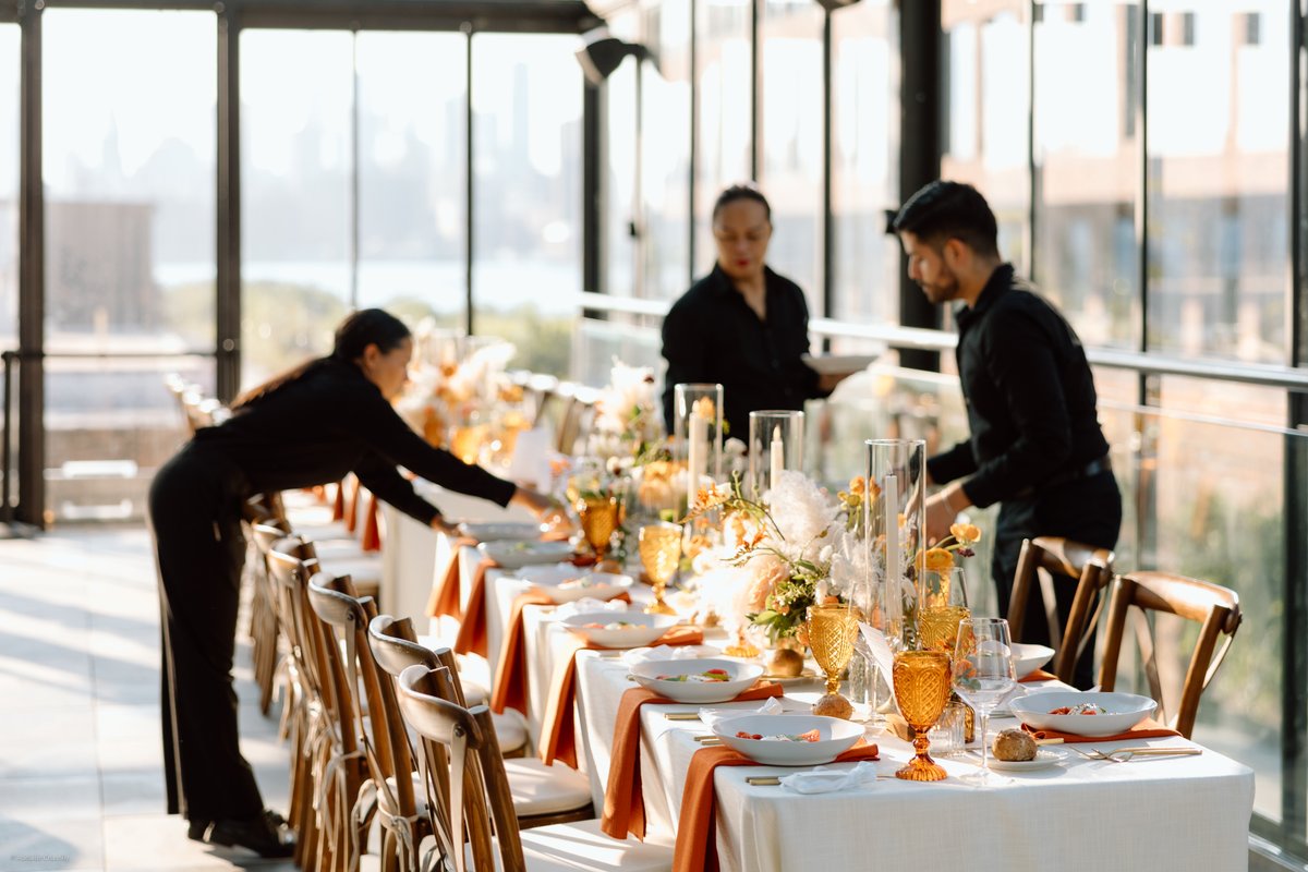 Three servers are diligently setting a long dining table for an event, with a blurred city skyline visible through large