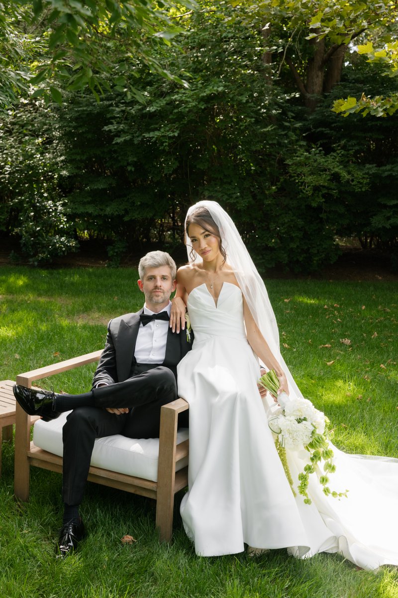 A newlywed couple poses elegantly outdoors on a sunny day. The groom sits on a wooden bench while the bride leans agains