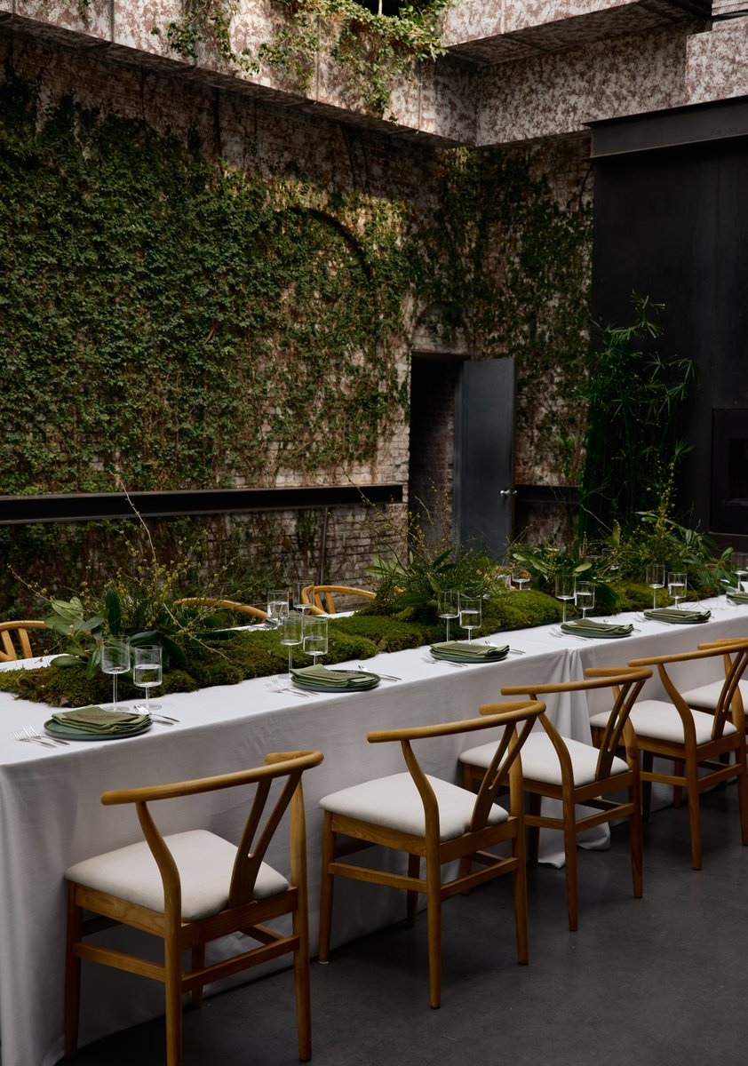 A long dining table is elegantly set for an event, featuring a lush, mossy and green foliage centerpiece. The setting is
