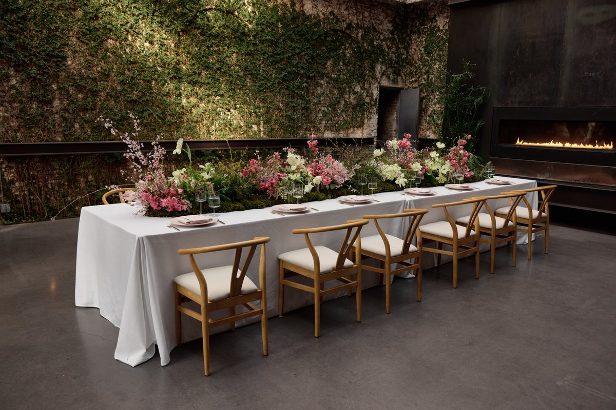 A beautifully set long dining table draped in a white tablecloth, featuring an elaborate floral centerpiece with pink an
