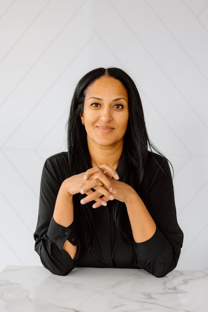 A professional portrait of a woman with long dark hair, wearing a black collared shirt, smiling gently at the camera. Sh