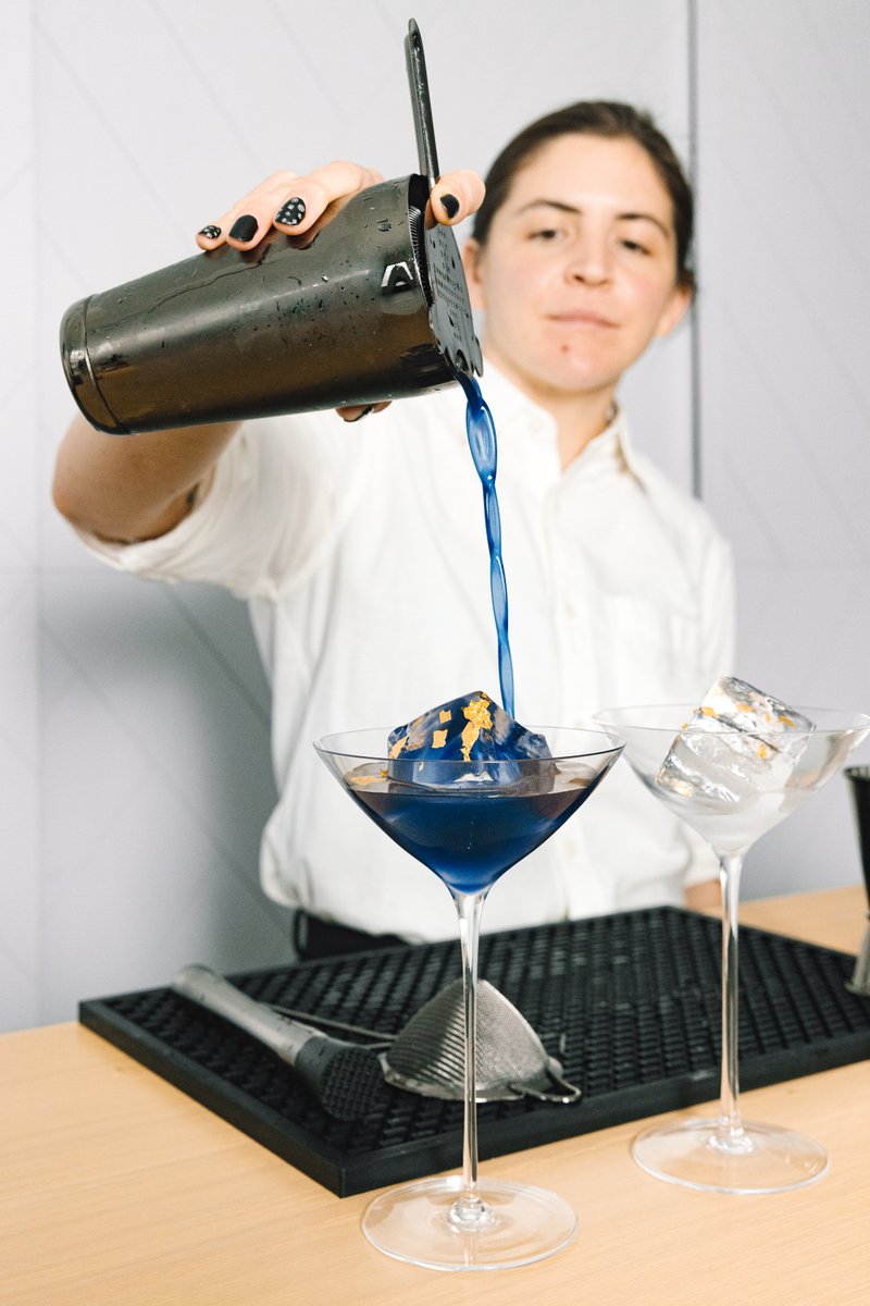 A bartender, wearing a white shirt, is expertly pouring a vibrant blue cocktail from a shaker into a martini glass. The