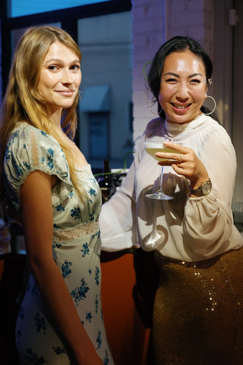 Two women are captured in a candid medium shot at an indoor event. The woman on the right smiles broadly while holding a
