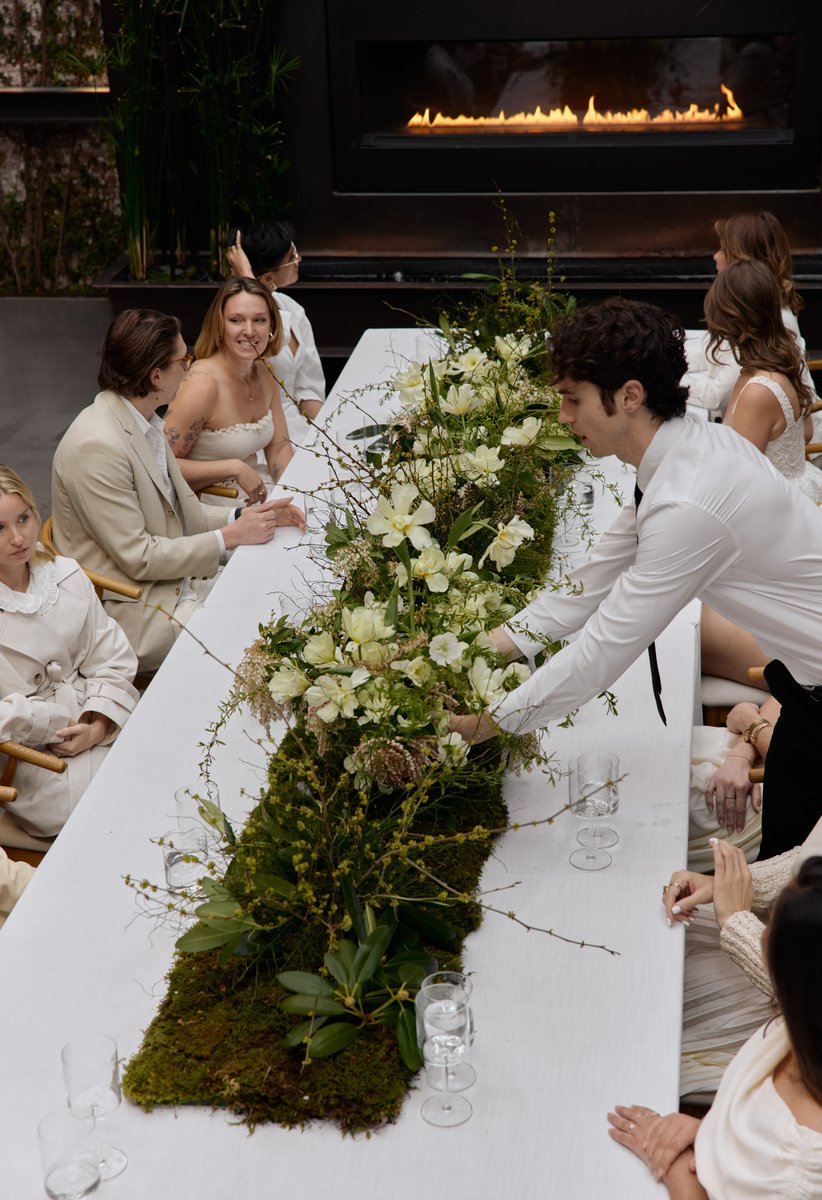 A server in a white shirt and black tie meticulously arranges a lush, long floral and moss centerpiece on a white dining