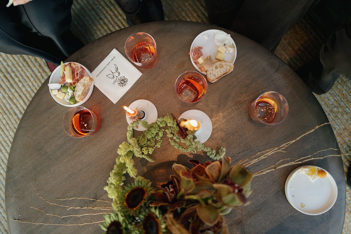 This overhead shot captures a round wooden table set with several cocktails, small appetizer plates, and floral decor. T