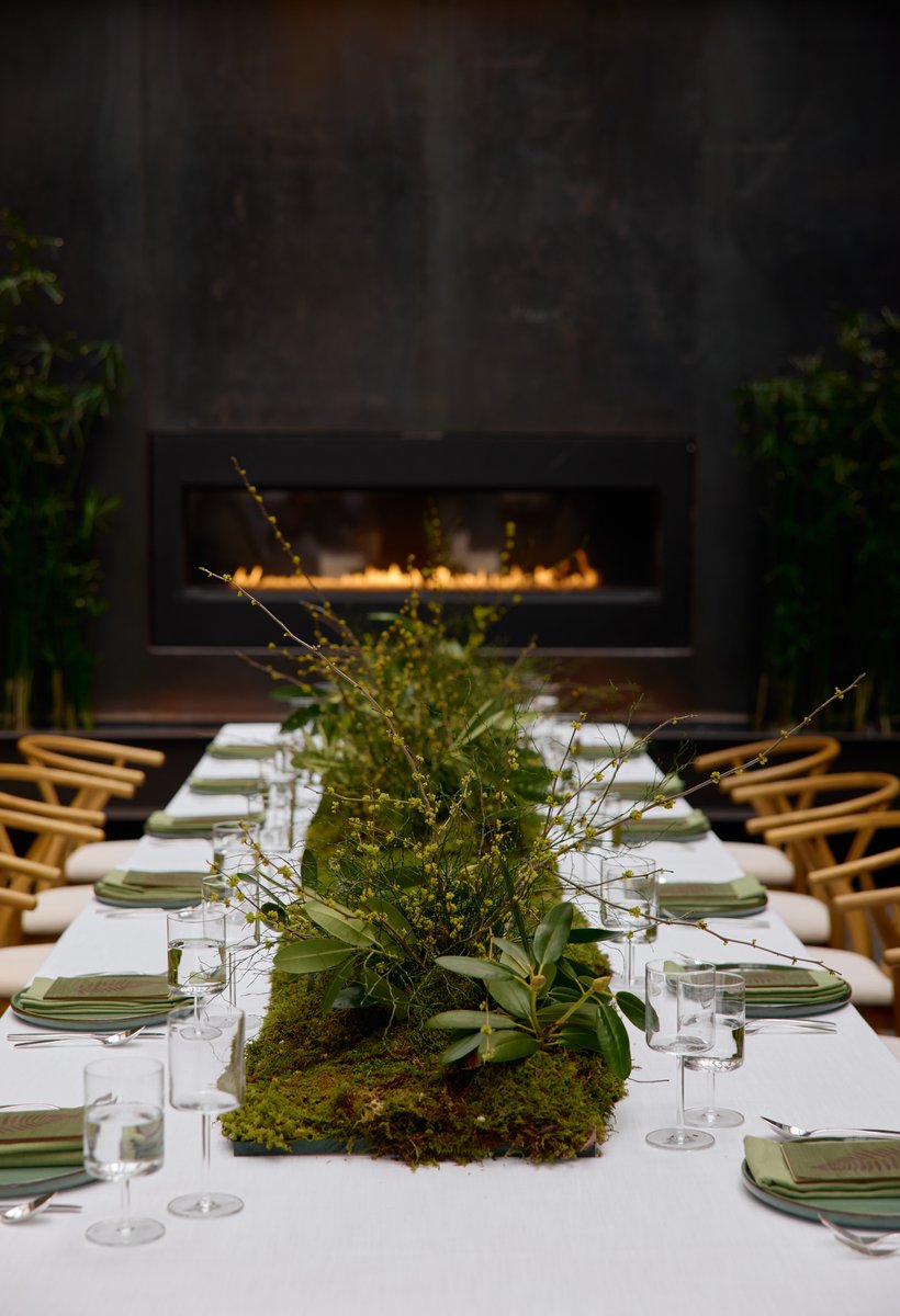 A beautifully set long dining table features a lush, mossy green centerpiece with various branches and foliage, creating