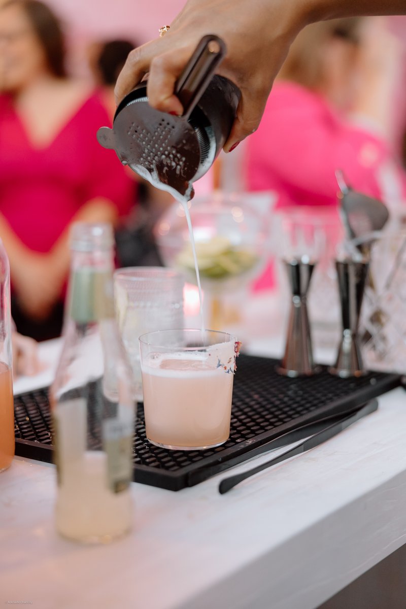 A close-up shot captures a bartender's hand pouring a light pink cocktail from a shaker into a glass with a floral-rimme